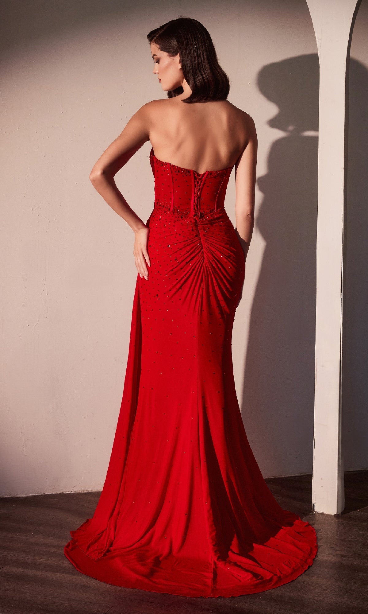A woman stands with her back to the camera, wearing the red Strapless Sweetheart Prom Gown Ladivine CD770, featuring a long corset design, ruched detailing, and a small train.